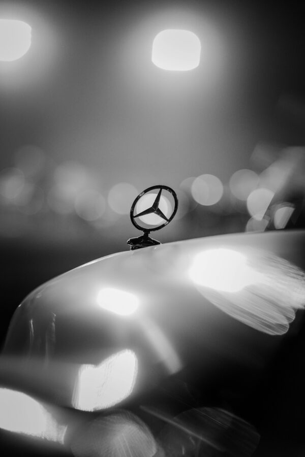 Black and white photo of a Mercedes emblem on car hood with bokeh lights, Ankara.