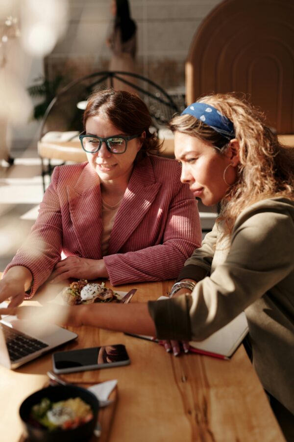 Two women engaged in teamwork at a café table, using a laptop in the sunlight.