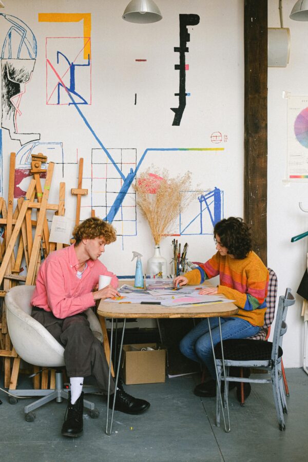 Full body side view of focused artists drawing pictures while sitting at table with art supplies in creative workshop with easels