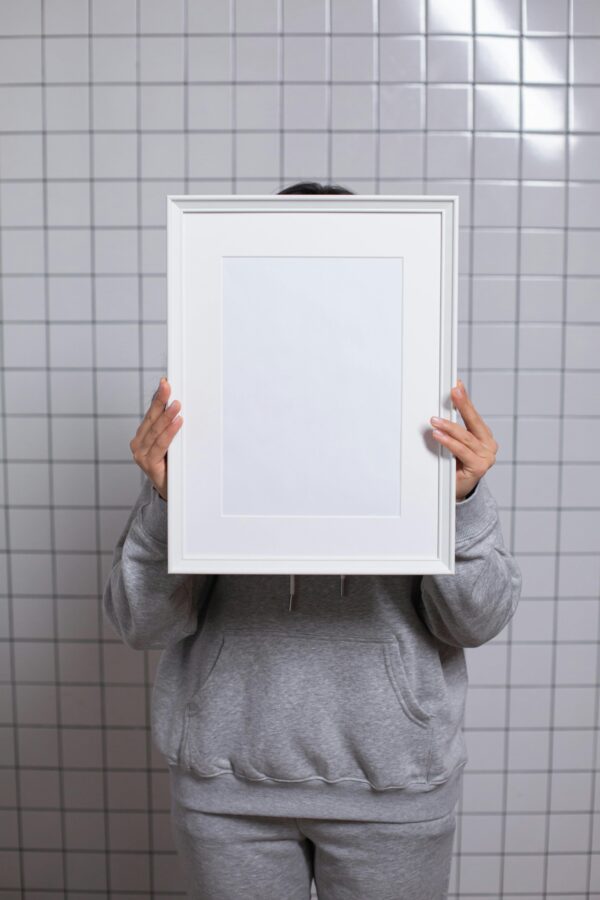 An unrecognizable individual holding an empty frame, standing against a tiled background, merged with a minimalist style.
