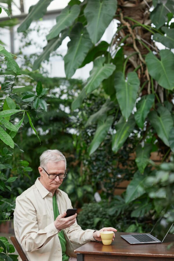 Elderly man using smartphone and laptop, enjoying coffee in a lush garden.